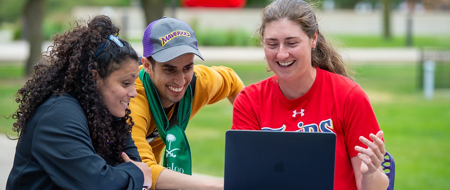 Three students outside on campus sitting down at a table looking at a laptop and smiling