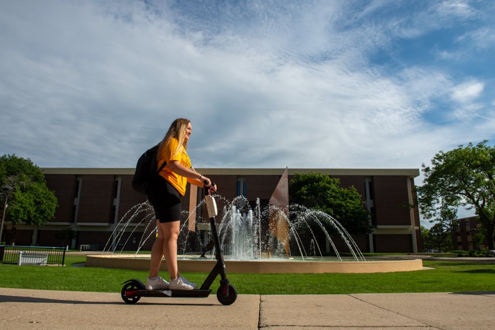 Student riding on a scooter in front of the MSU fountain.