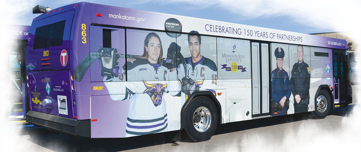 A City of Mankato bus outside of campus with the photo of two the Minnesota State Mankato hockey players celebrating 150 years of partnerships logo wrap