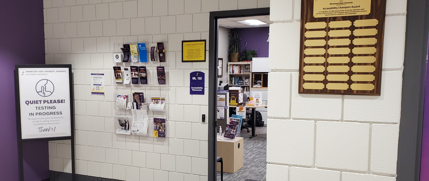 Hallway in front of an office labeled "Accessibility Resources," with a "Quiet Please! Testing in Progress" sign, brochure holders, and an "Accessibility Champion Award" plaque.