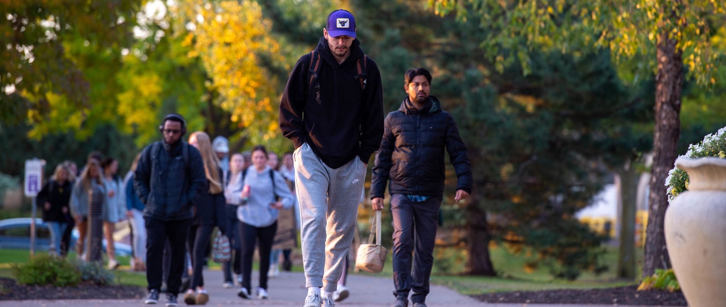 students walk on campus in the fall. Male student in the front wearing a Maverick purple hat