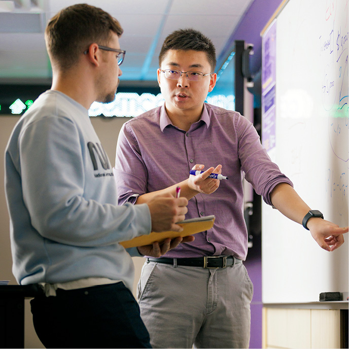 Professor of Finance in the finance lab with a student explaining a concept on the white board. Student taking notes