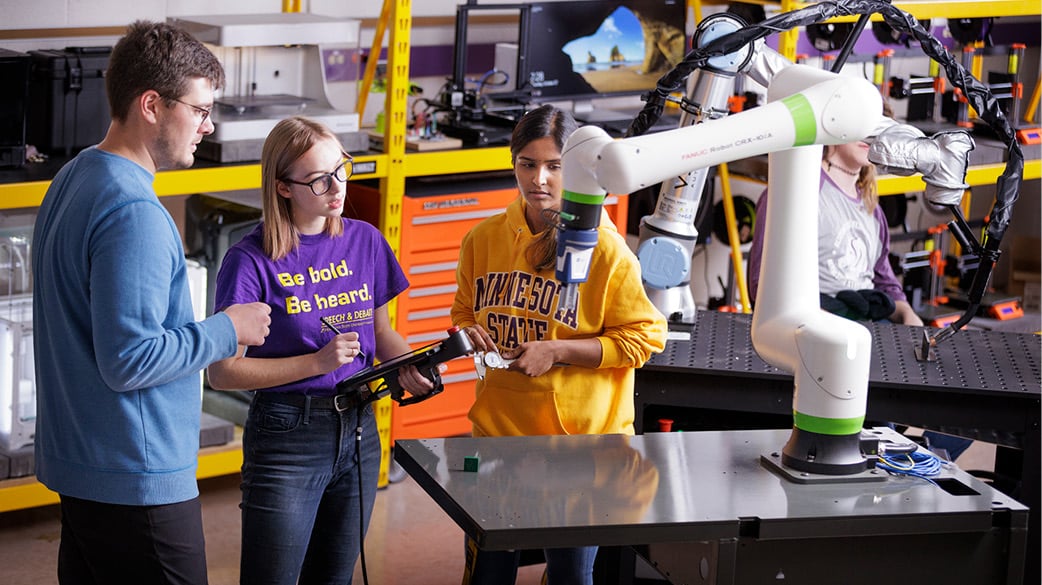 Two Minnesota State Mankato students working with a professor in a lab. Watching a robotic arm and taking notes on an ipdad.