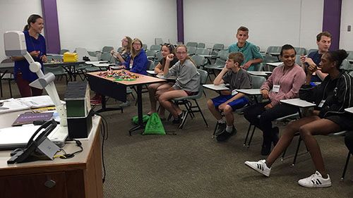 a large group of kids seated in a classroom listening to a volunteer