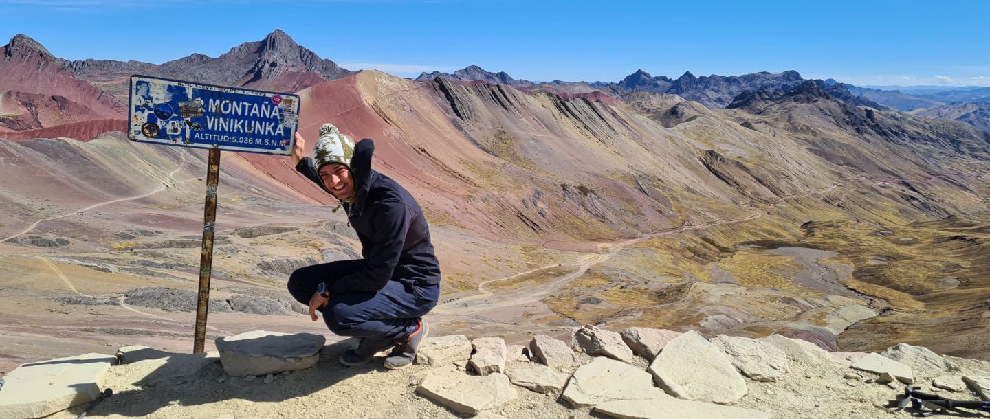 A student standing on top of Rainbow Mountain next to the sign posing with a smile and the mountain range in the background