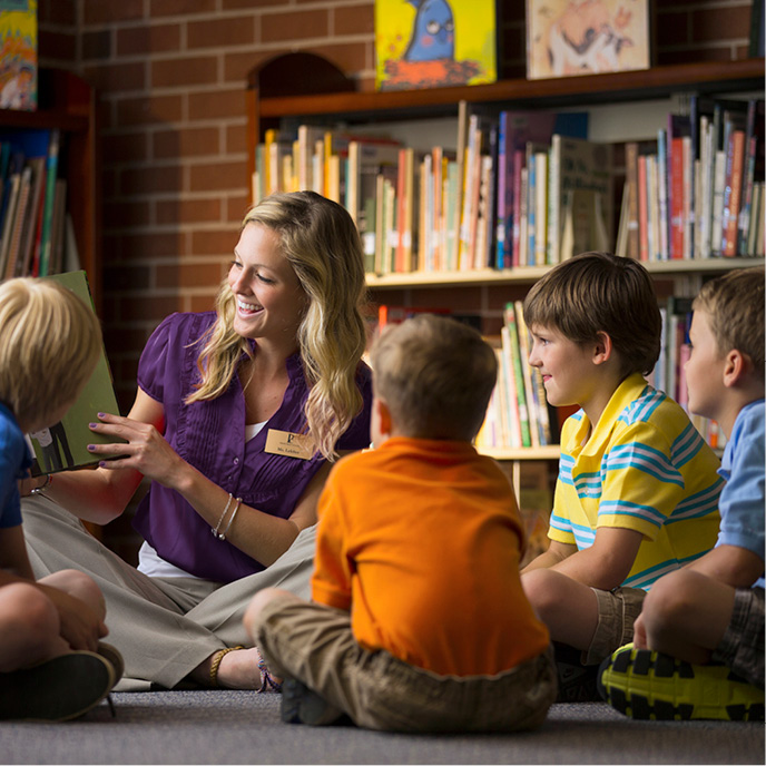 Student teacher sitting on the floor in a library reading a book to four elementary aged students