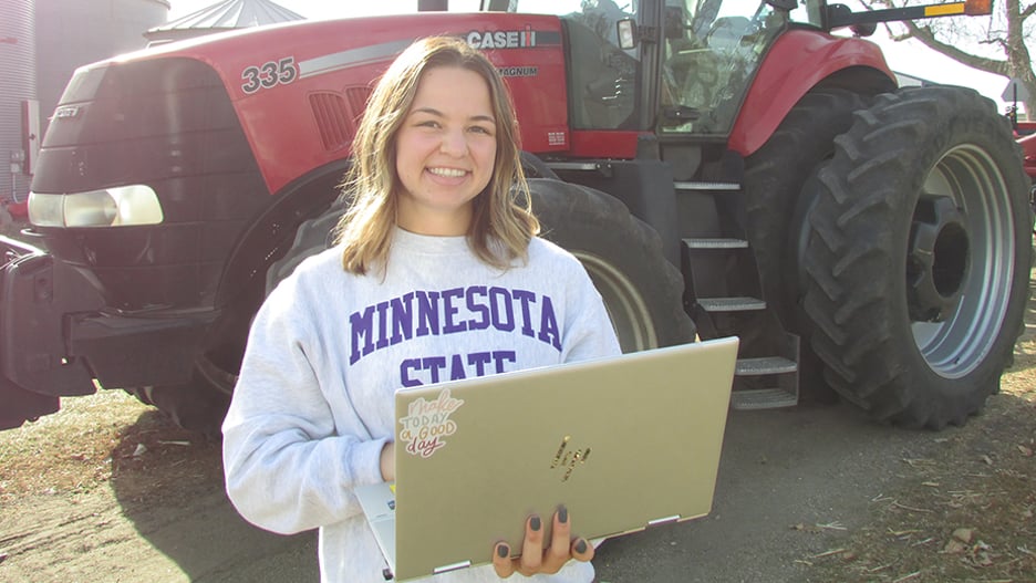 Maya Nelson posing with her laptop and a smile in front of a tracker on her family's Sleepy Eye farm