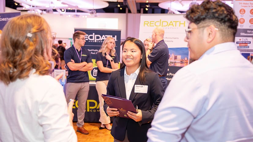 career fair with a student in a suit talking to two employers. The background is filled with students and employers